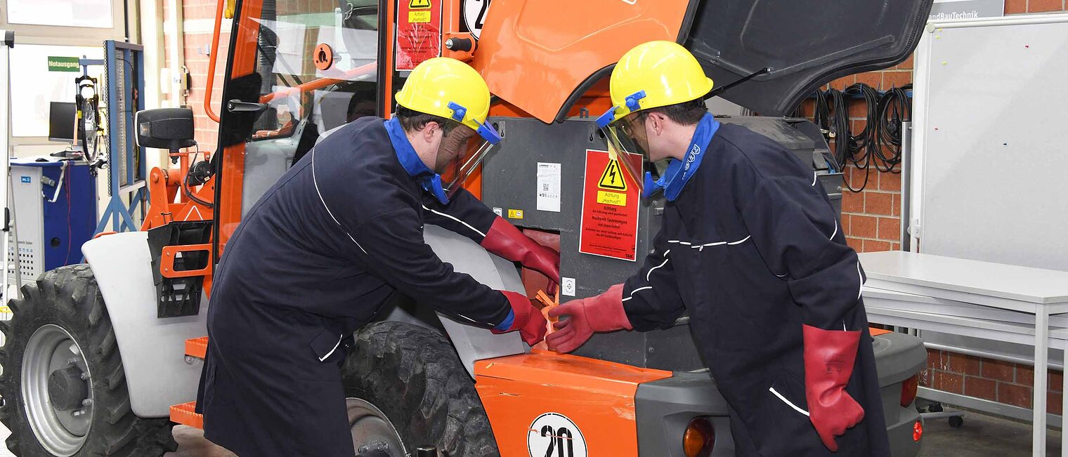 Fachkundige Person Hochvolt in der Land- und Baumaschinentechnik (3S) Zwei Männer mit Schutzkleidung führen Arbeiten im Hochvoltbereich an einem orangefarbenen Nutzfahrzeug in der Werkstatt durch
