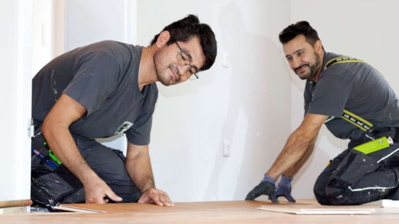 Zwei Männer knien in Arbeitskleidung auf dem Boden und verlegen Parkett