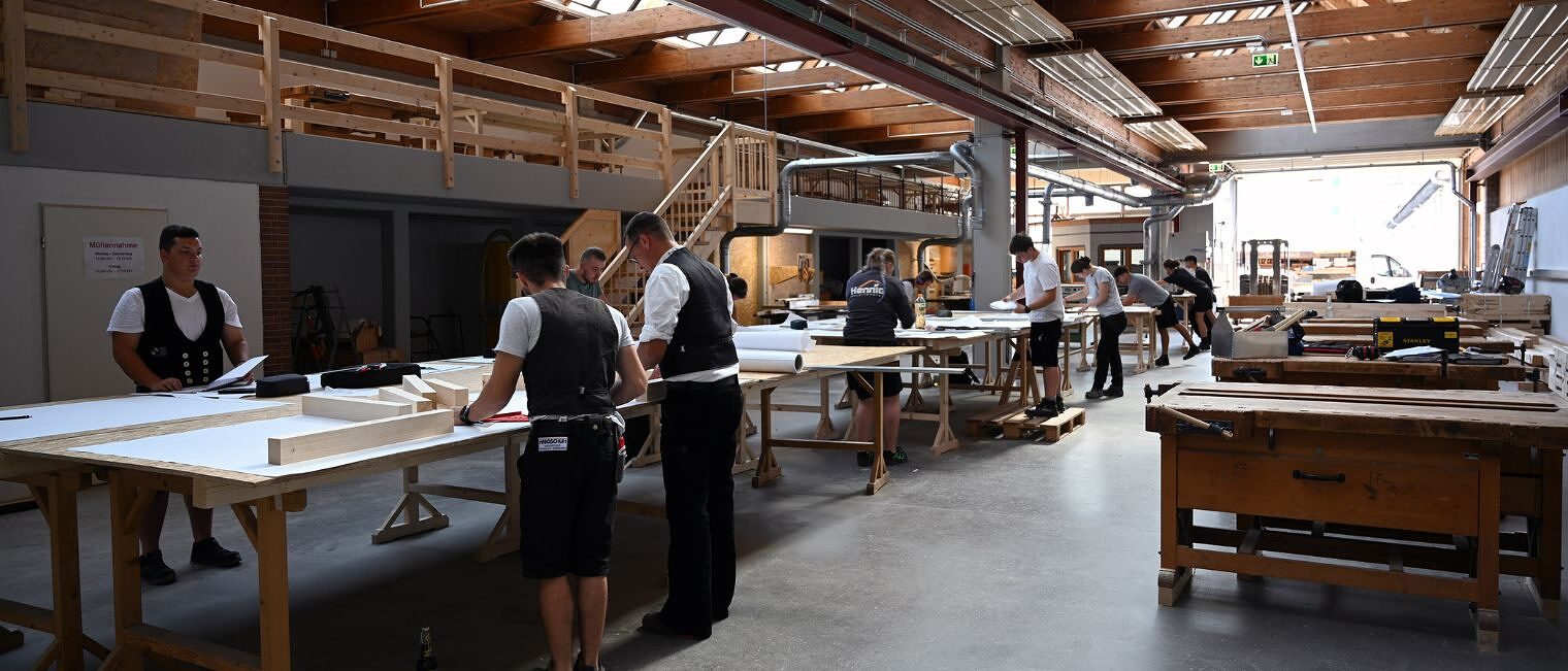 Zimmerer-Meisterschüler in Arbeitskleidung am Arbeiten an Tischen Große Halle mit mehreren Arbeitstischen, an denen Zimmerer-Meisterschüler in Arbeitskleidung und Kluft arbeiten