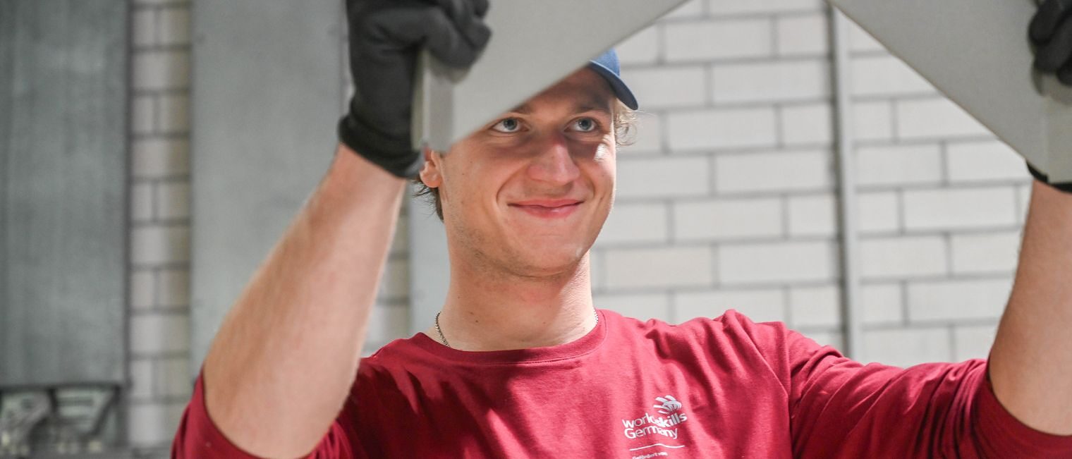 Junger Mann in rotem Oberteil (WorldSkills Logo) hält mit Arbeitshandschuhen ein Blech in die Höhe