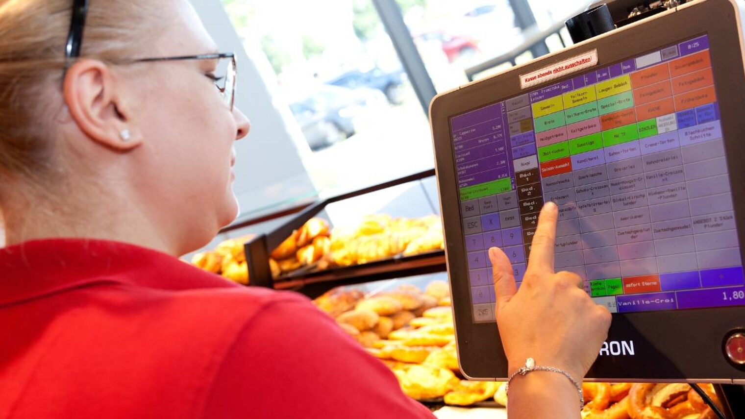 Eine Bäckereifachverkäuferin tippt hinter der Bäckertheke etwas auf einem Display der Kasse ein, trägt rotes T-Shirt und Zopf