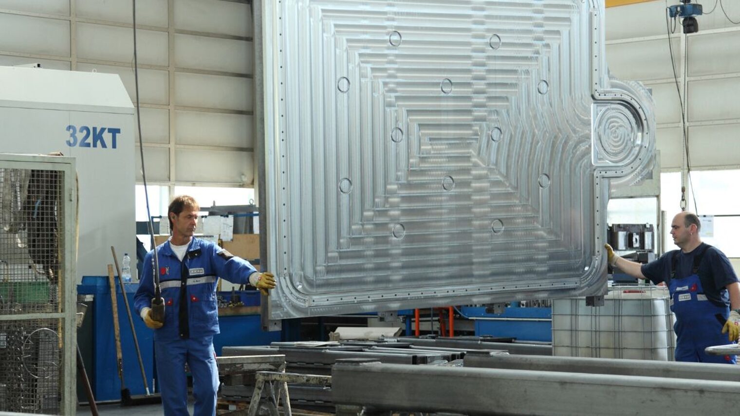 Zwei Männer in Blaumännern und Arbeitshandschuhen stehen an einer riesigen Metallplatte, die im Raum zwischen ihnen hängt