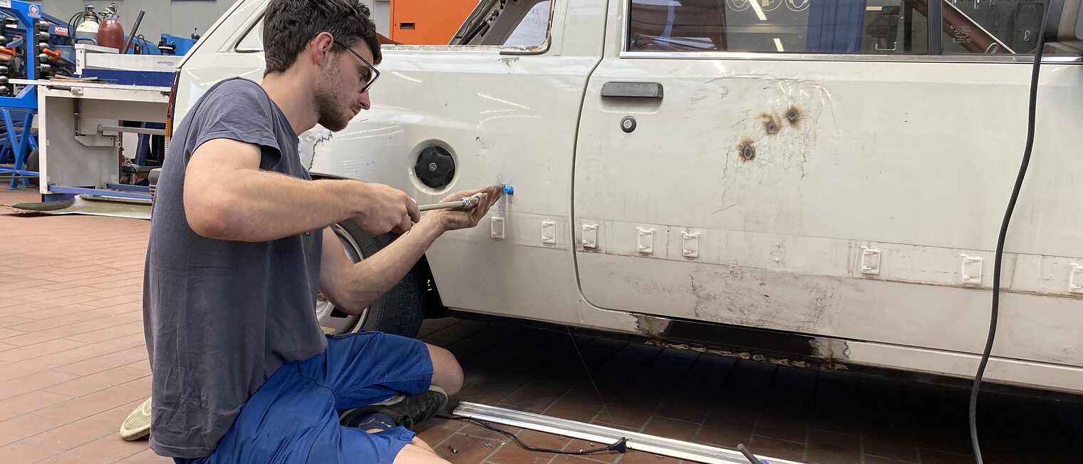 Ein Kursteilnehmer kniet in der Werkstatt und repariert die verrostete Seitenwand eines Autos mit Werkzeug.