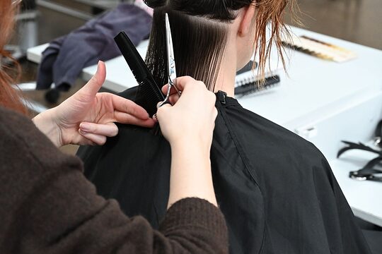 Situation von hinten fotografiert - eine Friseurin mit Kamm und Schere in der Hand schneidet einer sitzenden Dame die Haare
