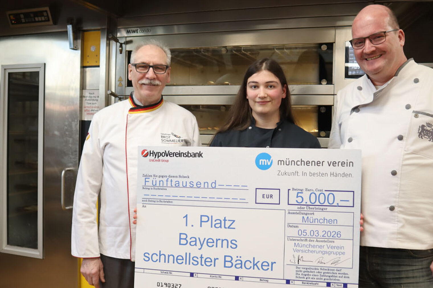 Drei Personen, in der Mitte Siegerin mit großem Symbolscheck